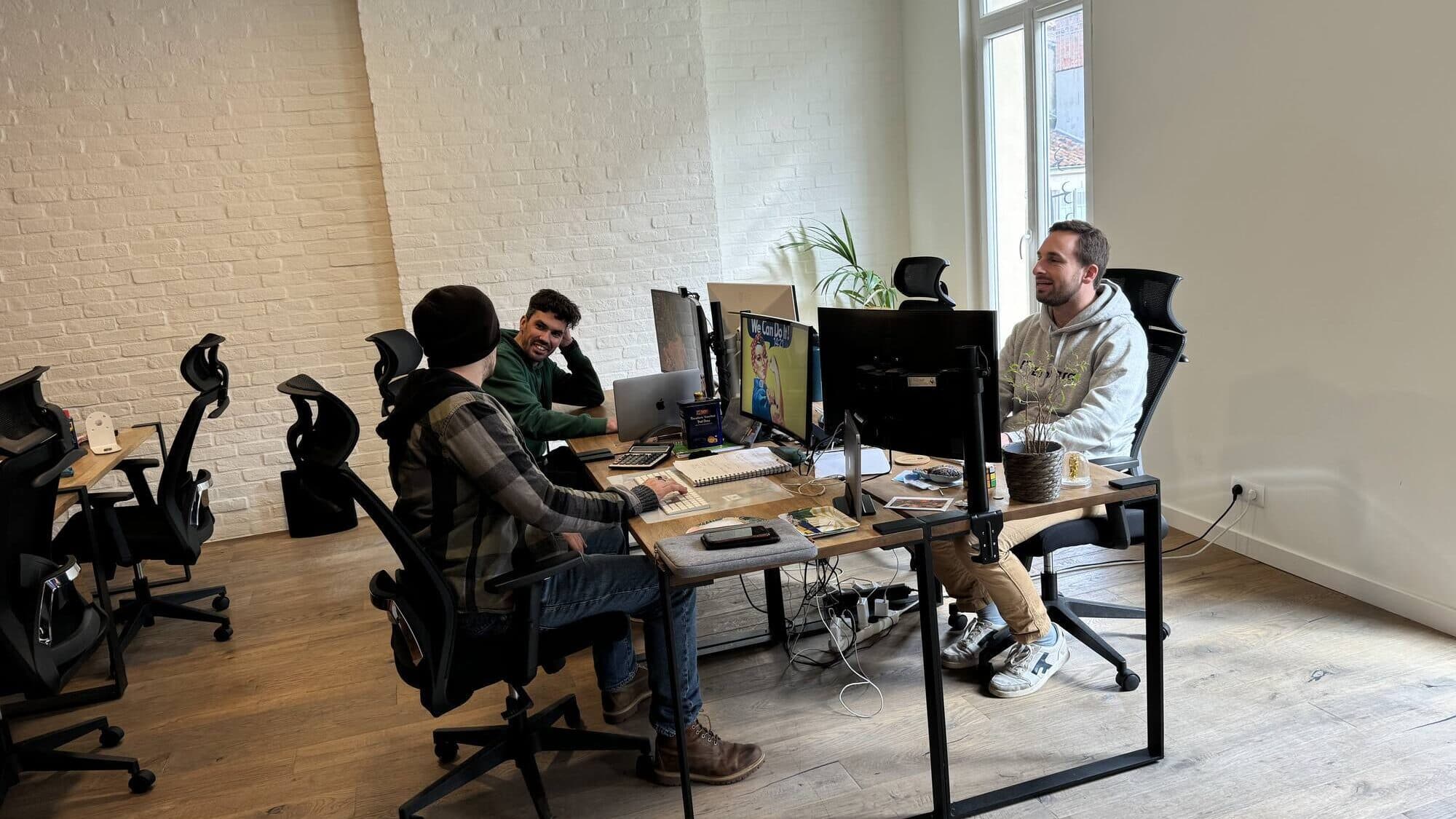 Trois personnes travaillent à leurs bureaux dans un open space aux murs de brique blanche